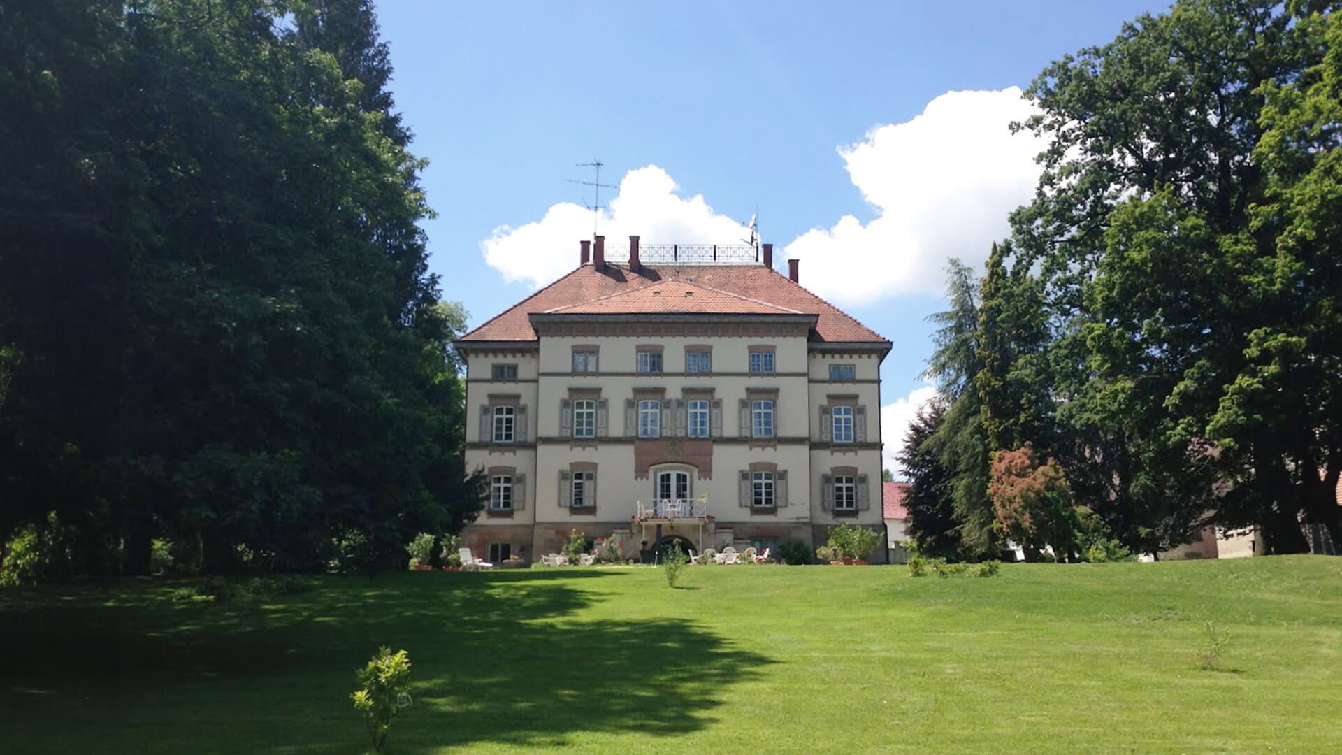 Das Schloss Schwendi steht eindrucksvoll vor einem strahlend blauen Himmel. Davor erstreckt sich eine große, grüne Graswiese, die dem historischen Gebäude eine offene und einladende Wirkung verleiht. Majestätische, große Bäume umrahmen das Schloss und schaffen eine harmonische Kulisse, die die idyllische Atmosphäre unterstreicht.
