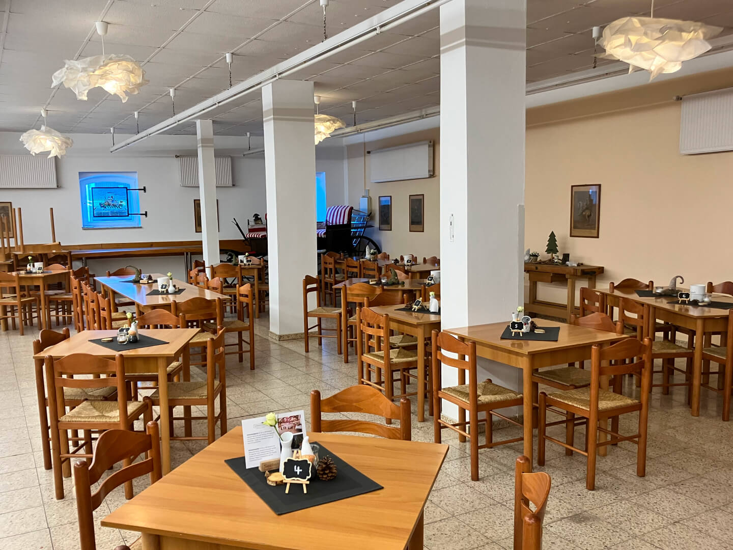 Der Innenraum wirkt hell und einladend mit seinen weißen Wänden, der weißen Decke und den elegant platzierten weißen Säulen. Eine warme Deckenbeleuchtung sorgt für eine gemütliche Atmosphäre. Zahlreiche Holztische und Holzstühle verleihen dem Raum eine natürliche, stilvolle Note und bilden einen harmonischen Kontrast zur weißen Umgebung.