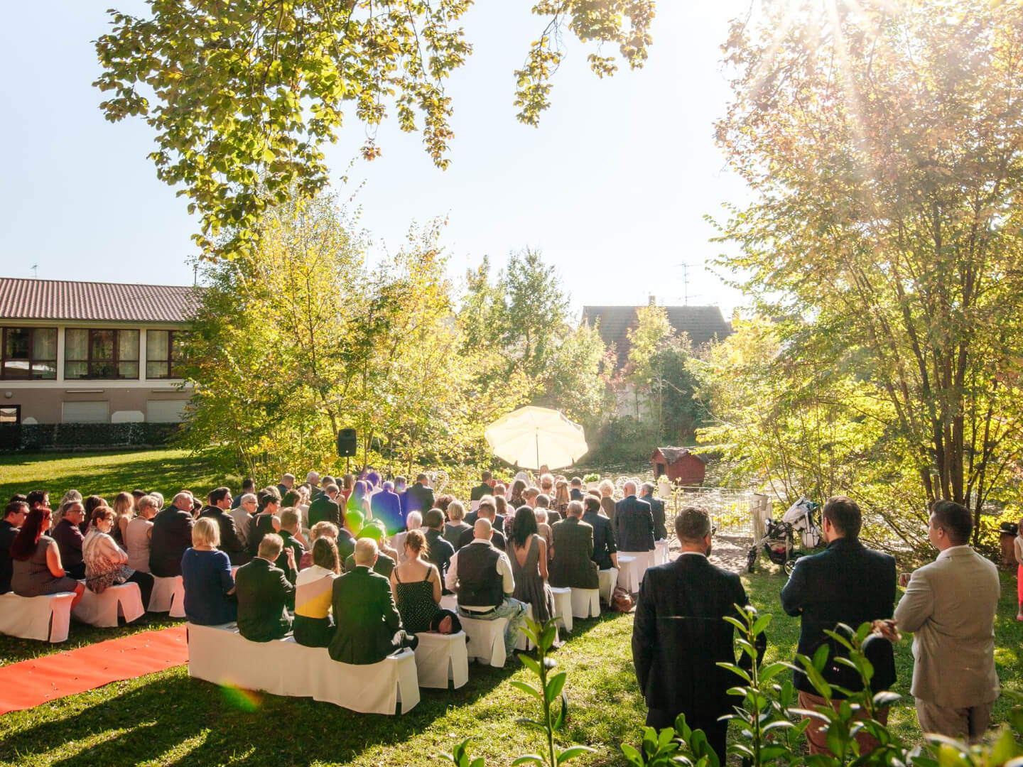 An einem sonnigen Tag in der Natur erstreckt sich eine malerische Landschaft mit zahlreichen Bäumen, üppigen Pflanzen und einem ruhigen See. Im Hintergrund sind einige Häuser zu sehen, die sich harmonisch in die Umgebung einfügen. Vor dem See versammelt sich eine feierliche Gesellschaft, die die idyllische Kulisse für ein besonderes Ereignis genießt.