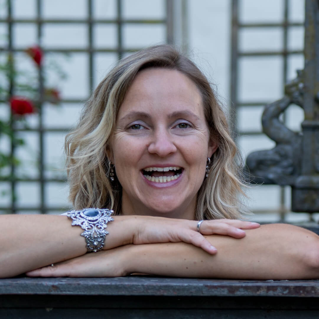 Eine blonde Frau stützt ihre Arme auf einer Mauer ab und blickt mit einem fröhlichen Lächeln darüber. Ihr Blick ist entspannt, und sie lacht herzlich in die Kamera, was eine warme und einladende Atmosphäre schafft. Die Szene wirkt spontan und lebendig, mit einem Hauch von Freude und Unbeschwertheit.